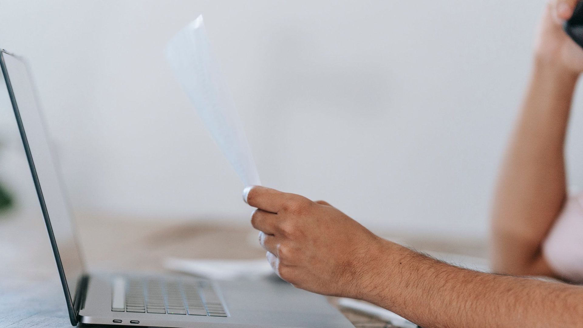 Homem a rever um documento e a falar ao telemóvel, sentado a uma secretária com um portátil