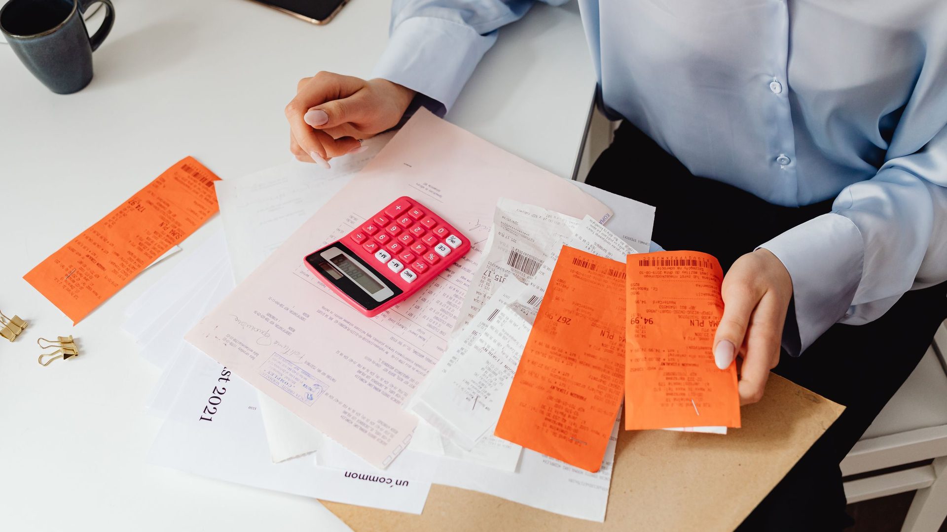 Mãos de uma pessoa separando recibos coloridos e usando uma calculadora rosa sobre uma mesa